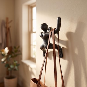 May include: A dark, textured metal wall hook shaped like a human figure in a climbing pose. The figure is mounted to the wall with two visible screws, one at the head and one at the foot, and is currently holding the brown leather straps of a bag.