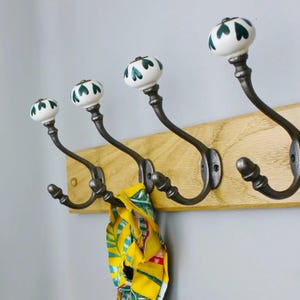 May include: A wall-mounted coat rack featuring a light-coloured wooden board with four dark metal hooks. Each hook is topped with a white ceramic knob decorated with a green heart pattern. A yellow patterned fabric item hangs from one hook.