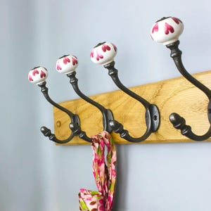 May include: A wall-mounted coat rack featuring a light-coloured wooden board with four dark metal hooks. Each hook is topped with a white ceramic knob decorated with a repeating pattern of small, dark pink heart-shaped petals.