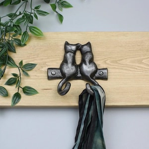 May include: A dark grey metal wall hook featuring two cat silhouettes sitting side by side, mounted on a light brown wooden board. The hook is secured with two visible screws, and a dark patterned fabric scarf hangs from one of the cat tails.