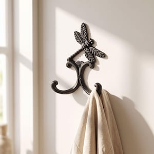 May include: A dark brown, cast iron wall hook featuring a dragonfly motif at the top and two curved hooks at the bottom. The metal hook is mounted to a wall and holds a piece of light beige fabric draped over one of the lower curved arms.