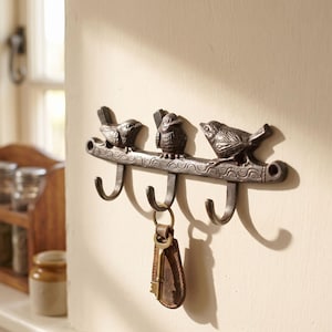 May include: A dark brown metal wall hook rack featuring three sculpted birds perched on a horizontal bar with a wavy pattern. The rack has three curved hooks, with a single metal key on a brown leather fob hanging from the centre hook.