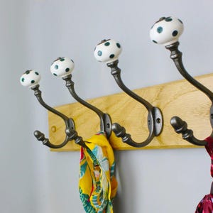 May include: A wall-mounted coat rack featuring a light-coloured wooden board with four dark metal hooks. Each hook is topped with a white ceramic knob decorated with small green polka dots. A yellow patterned scarf hangs from the second hook.