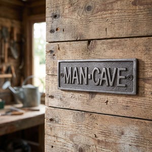 May include: A rectangular, dark grey metal sign with a hammered texture background and raised, smooth lettering that reads MAN-CAVE. The sign is secured to a wooden surface with two visible screws, one on the far left and one on the far right.
