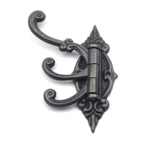 May include: A dark grey metal wall hook featuring three curved arms with rounded tips. The base plate has an elongated, ornate shape with decorative scrollwork and two screw holes for mounting, providing a functional design for hanging items.