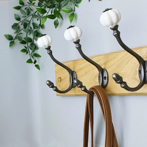 May include: A wall-mounted coat rack featuring three dark metal hooks attached to a light-coloured wooden board. Each hook is topped with a white, pumpkin-shaped ceramic knob. A brown leather strap hangs from the centre hook of the rack.