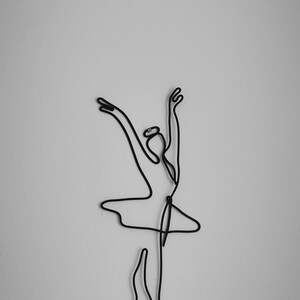 May include: A black wire sculpture of a ballerina in a pose with arms raised above her head.