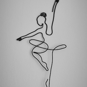 May include: A black wire sculpture of a ballerina in a pose with one leg extended and the other leg bent at the knee. The ballerina's arms are outstretched, one above her head and the other to the side.