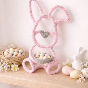May include: A pink crochet Easter bunny ornament with a grey heart detail and a small basket filled with pastel eggs. A wicker basket with eggs and a white ceramic bunny are also visible.