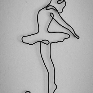 May include: A black wire sculpture of a ballerina in a pose with her arms raised above her head and her legs bent at the knees. The sculpture is mounted on a white wall.