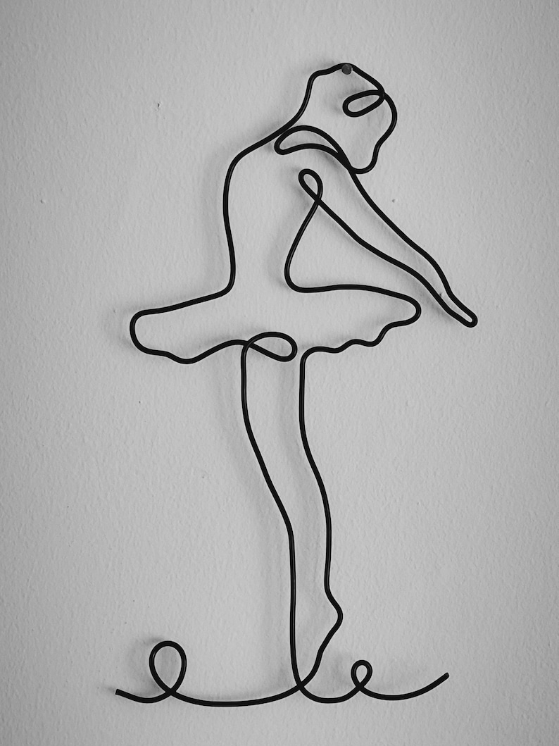 May include: A black wire sculpture of a ballerina in a pose with her arms raised above her head and her legs bent at the knees. The sculpture is mounted on a white wall.