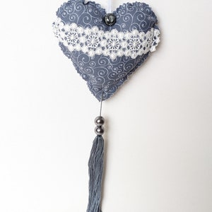 May include: A grey heart-shaped fabric ornament with white lace trim and a grey tassel hanging from it. The heart has a swirl pattern and a grey button in the centre.