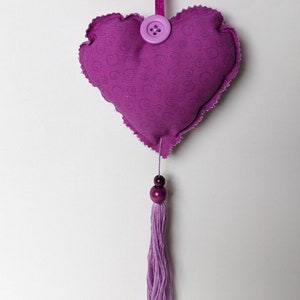May include: A purple heart-shaped decoration with a scalloped edge, a button, and a tassel. The heart is made of fabric with a swirl pattern. A purple ribbon is attached for hanging. The tassel is made of purple thread and has two wooden beads.