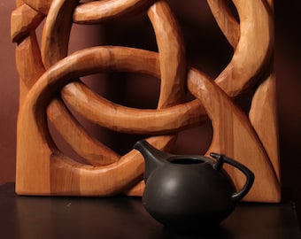 Könnte beinhalten: Eine Holz-Wandskulptur mit einem komplexen, ineinander verschlungenen Design, im Vordergrund eine schwarze Keramik-Teekanne.