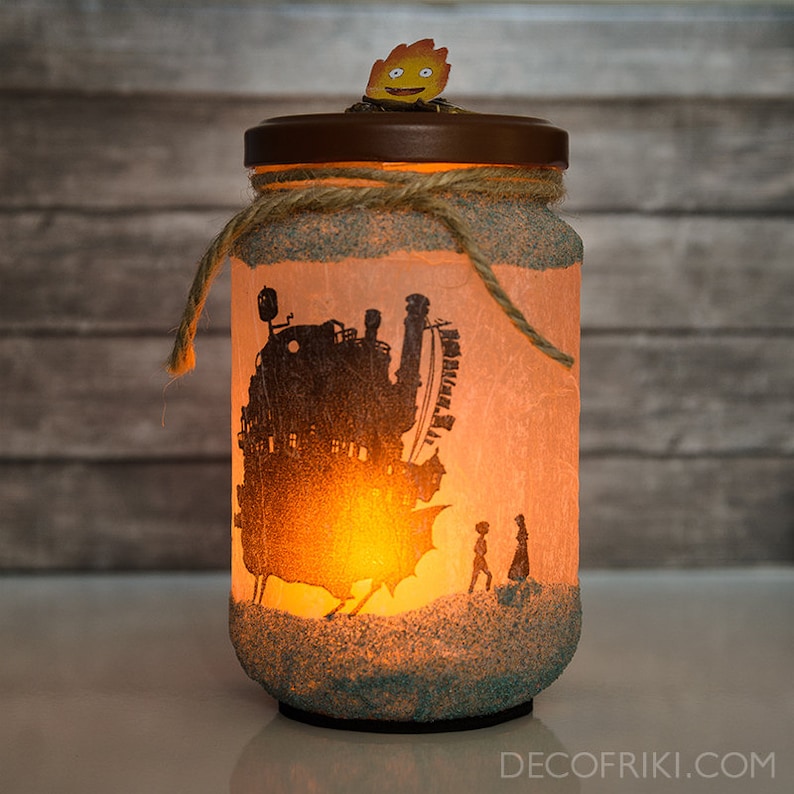 K&ouml;nnte beinhalten: Ein Glasgef&auml;&szlig; mit einem hellbraunen Deckel und einer Silhouette eines fliegenden Hauses, zu dem eine kleine Figur geht. Das Gef&auml;&szlig; ist mit braunem Garn und hellblauem Glitzer am Boden verziert. Das Gef&auml;&szlig; wird von innen mit einem warmen gelben Licht beleuchtet.