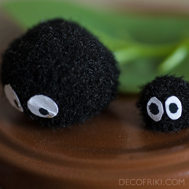 May include: Two small, fuzzy black creatures with large, white-rimmed eyes. The larger one is slightly out of focus, while the smaller one is in sharp focus. The creatures are set against a blurred green background.