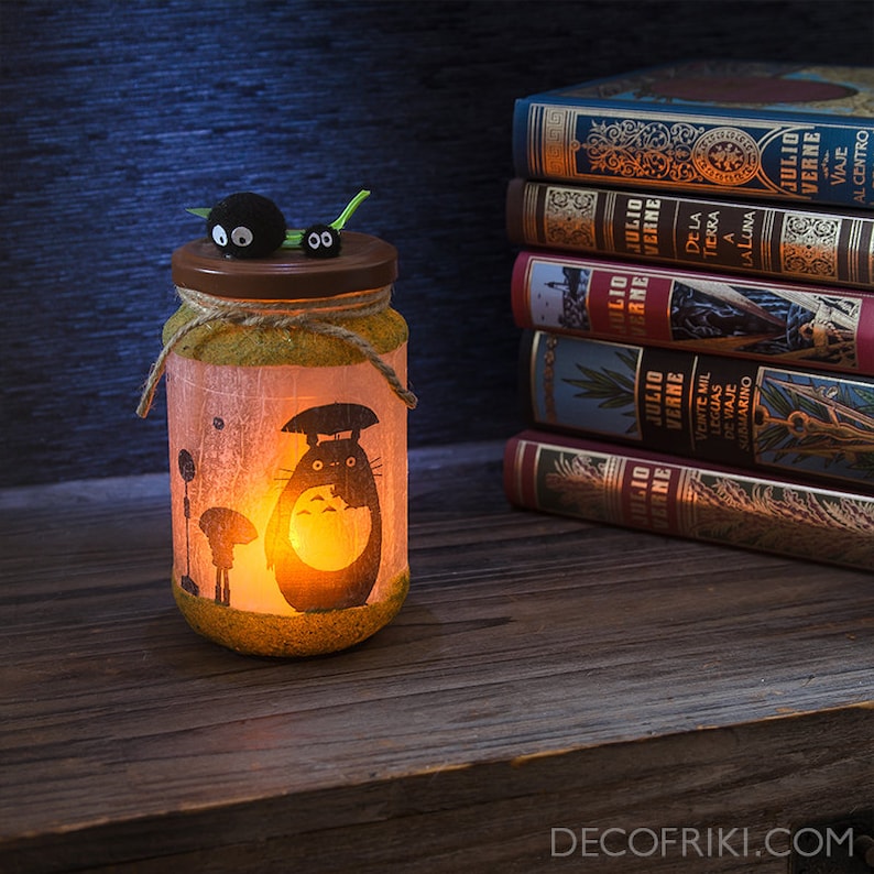 May include: A decorative glass jar with a Totoro design, lit from within, sits on a wooden surface. The jar is adorned with twine and a lid with small, black, cartoon-like figures. A stack of books with gold lettering is in the background.
