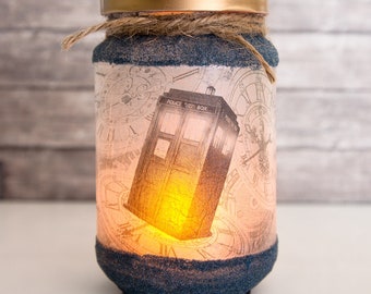 May include: A glass jar with a blue and white design featuring a TARDIS, the iconic time machine from the Doctor Who television series. The jar is decorated with a brown twine bow and has a gold lid. The jar is lit from within, creating a warm glow.