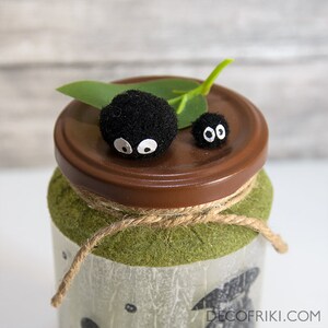 May include: A decorative jar with a brown lid, adorned with two black, fuzzy creatures with white eyes. The jar is wrapped with twine and has a green textured base. A green leaf adds a natural touch to the design.