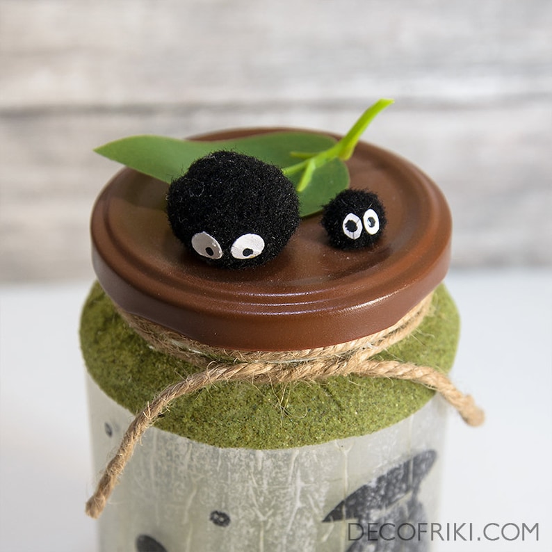 May include: A decorative jar with a brown lid, adorned with two black, fuzzy creatures with white eyes. The jar is wrapped with twine and has a green textured base. A green leaf adds a natural touch to the design.