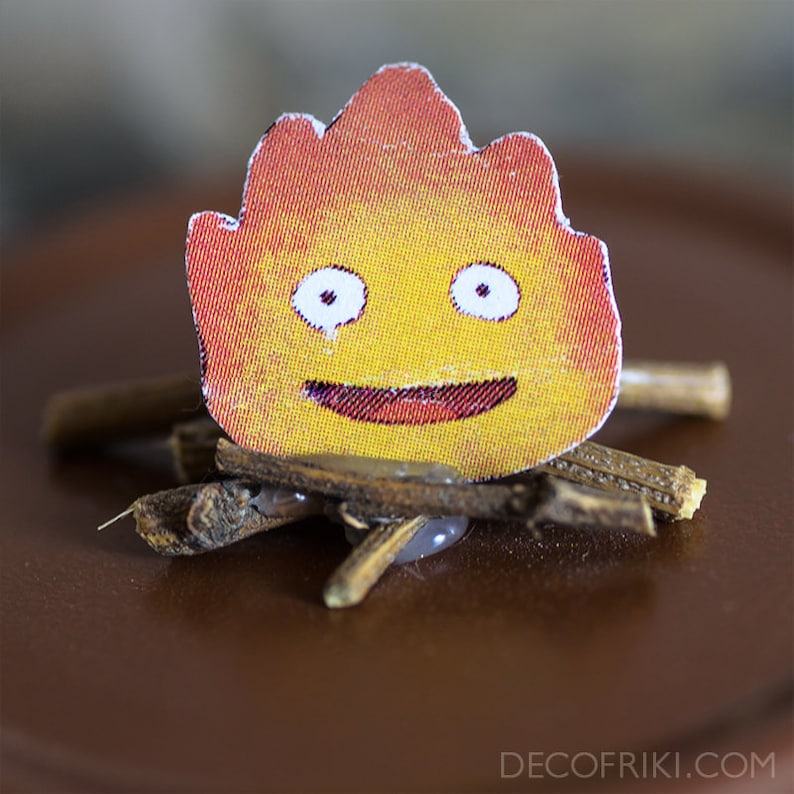 K&ouml;nnte beinhalten: Eine l&auml;chelnde, gelbe und orangefarbene Feuerspirit-Figur mit wei&szlig;en Augen, die auf einem Bett aus braunen Zweigen sitzt.