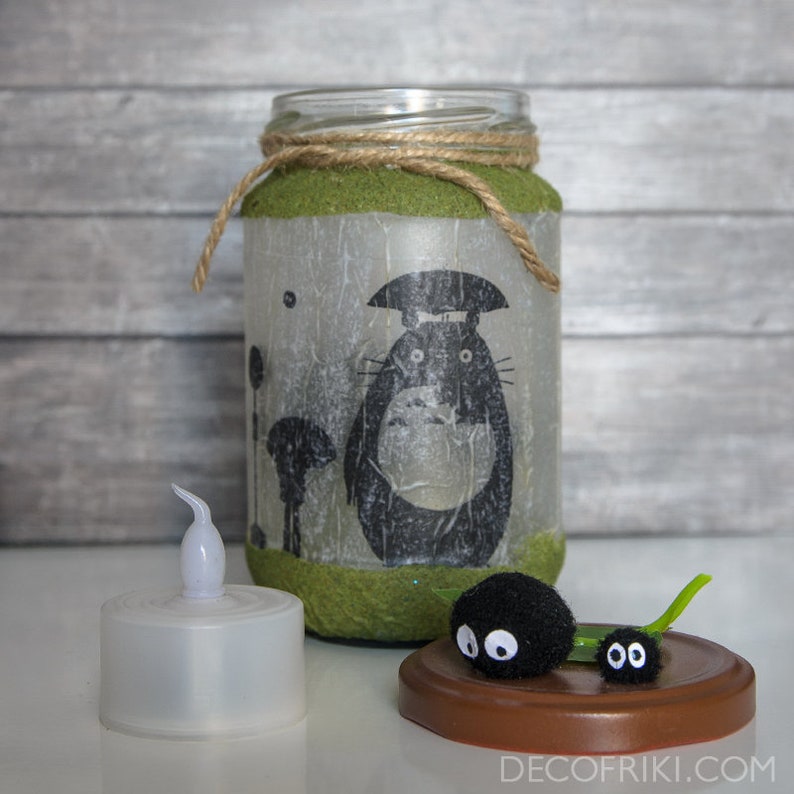May include: A glass jar lantern featuring a black Totoro design, wrapped with twine and green material. A small white LED candle and two black soot sprites sit nearby on a brown base. The lantern is a decorative item.