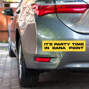 May include: A gray car's rear bumper with a yellow rectangular sticker. The sticker has black text that reads "IT'S PARTY TIME IN DANA POINT". The car's taillights are red.