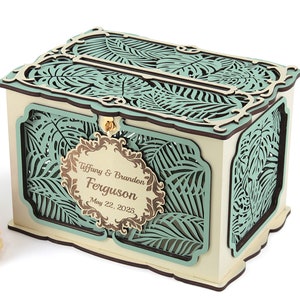 May include: A wooden wedding card box with a teal green and brown tropical leaf design. The box has a slot on the top for cards and a hinged lid. The box is personalized with the names "Tiffany & Brandon Ferguson" and the date "May 22, 2025".