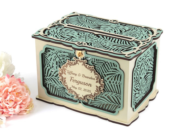 May include: A wooden wedding card box with a teal green and brown tropical leaf design. The box has a slot on the top for cards and a hinged lid. The box is personalized with the names "Tiffany & Brandon Ferguson" and the date "May 22, 2025".