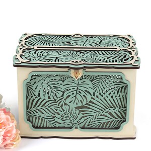 May include: A wooden box with a teal tropical leaf design. The box has a slot on the top for inserting cards or envelopes.