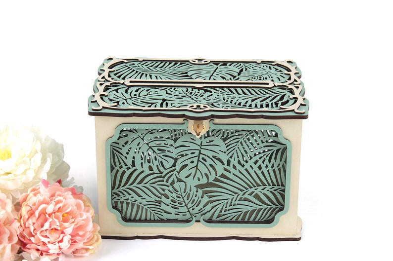 May include: A wooden box with a teal tropical leaf design. The box has a slot on the top for inserting cards or envelopes.