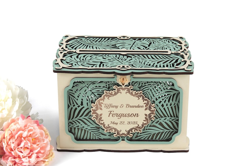 May include: A wooden wedding card box with a teal and brown tropical leaf design. The box has a slot for cards and a personalized label that reads "Tiffany & Brandon Ferguson May 22, 2025."