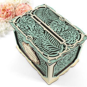 May include: A teal and brown wooden box with a cutout design of tropical leaves. The box has a slot on the top for cards or envelopes. The box is perfect for a wedding or other special occasion.
