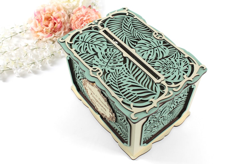 May include: A teal and brown wooden box with a cutout design of tropical leaves. The box has a slot on the top for cards or envelopes. The box is perfect for a wedding or other special occasion.