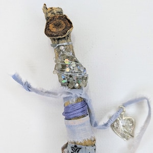 May include: A handcrafted wooden wand with a natural bark finish. The wand is adorned with silver wire, iridescent beads, and wrapped with blue and patterned fabric. A silver heart charm hangs from a ribbon.