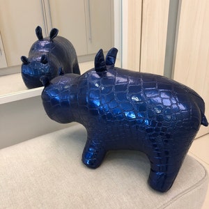 May include: Blue plush rhinoceros toy with a crocodile skin texture. The toy is sitting on a light beige fabric surface.
