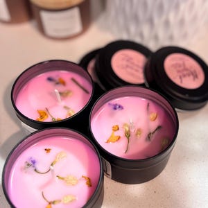 Puede incluir: Velas rosas en latas negras, adornadas con flores secas. Las velas tienen mechas blancas y están rodeadas de otras velas con tapas negras. El tema es la fragancia y la decoración del hogar.