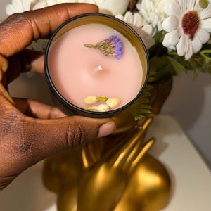 以下が含まれることがあります： 黒い缶に入った小さな丸いキャンドル。ピンク色のワックスはドライフラワーで飾られています。キャンドルは手で持たれており、背景には金色の装飾品と白い花のブーケがあります。キャンドルはセットの一部です。