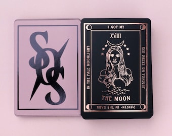 May include: Two tarot cards, one with a black and white illustration of a stylized letter "S" on a light pink background, and the other with a black background and a rose gold illustration of a woman with long hair and a crescent moon above her head. The text on the card reads "I got my XVIII THE MOON DANCIN' IN THE DARK RED DRESS ON TONIGHT"