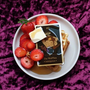 May include: A white plate with a waffle topped with butter and syrup, surrounded by sliced strawberries. A card with the text "The Waffles + Indulgence" and a number "40" is on top of the waffle. The plate is on a purple velvet surface.