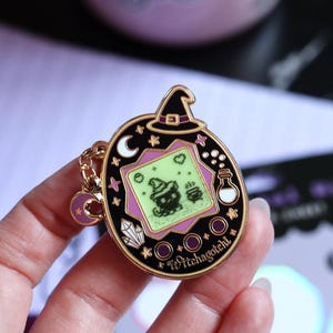May include: A black and gold metal enamel pin shaped like a digital pet device, featuring a witch hat on top, a screen with a cat and cauldron graphic, and the text Witchagotchi. A small purple crescent moon charm hangs from a gold chain on the side.