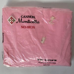 May include: A pink Cannon Monticello no-iron fitted bottom sheet packaged in clear plastic. The label indicates it is one sheet for a 54 by 75 inch mattress, made of 50 percent cotton and 50 percent polyester, with elastic edges, made in the U.S.A.
