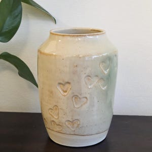 May include: A cream-colored ceramic vase with a cylindrical shape and a slightly flared rim. The vase features a series of embossed heart shapes across its surface. The base of the vase is a lighter shade of white.