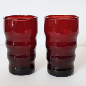 May include: Two ruby red drinking glasses. Each glass has a wavy, textured design and a dark base. The glasses are cylindrical with a slightly flared rim. The glasses are made of a transparent material.