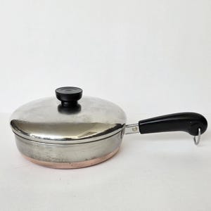 May include: A stainless steel skillet with a copper-colored base and a matching lid. The pan features a black heat-resistant handle with a small metal hanging loop at the end and a black knob on the center of the lid for easy lifting.