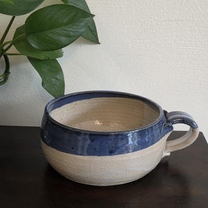 May include: A wide, round ceramic bowl with a single handle, featuring a two-tone design with a glossy dark blue glaze on the upper portion and a speckled, off-white matte finish on the lower half, creating a distinct contrast in texture and color.