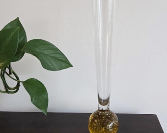May include: A tall, slender clear glass bud vase featuring a spherical base filled with yellow-tinted glass and numerous small air bubbles. The vase has a long, narrow neck that flares slightly at the top, standing upright on a flat surface.