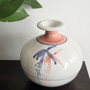 May include: A white ceramic vase with a round body and a narrow neck. The vase features a coral-colored band around the neck and blue and coral abstract floral designs. The vase is displayed on a dark surface.