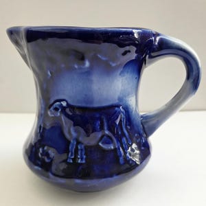 May include: A cobalt blue ceramic pitcher with a handle and spout. The pitcher features a raised relief of a cow on one side. The glaze has a glossy finish. The pitcher is approximately 5 inches tall.