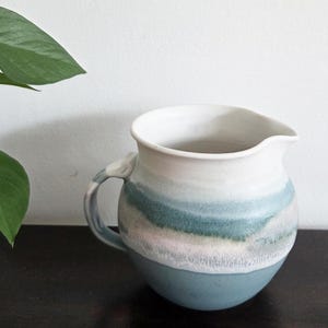 May include: A ceramic pitcher with a rounded base and a spout. The pitcher features a color gradient, transitioning from a teal base to white at the top, with bands of blue and pink in between. It has a curved handle with a decorative element.